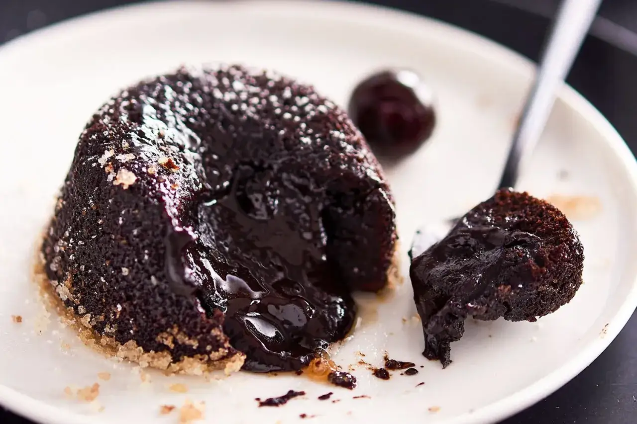Tortino al cioccolato con cuore fuso, spolverato di zucchero a velo, accanto a una ciliegia scura.