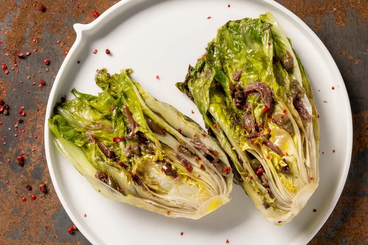 Scarola in padella con acciughe, olive e pepe rosa su piatto bianco.