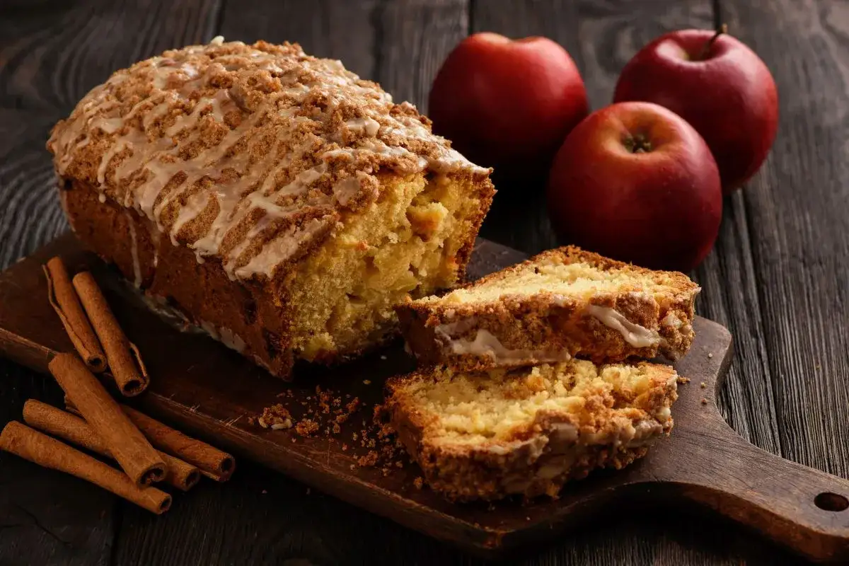 Plumcake alle mele con glassa e crumble, mele rosse e cannella su tagliere di legno.