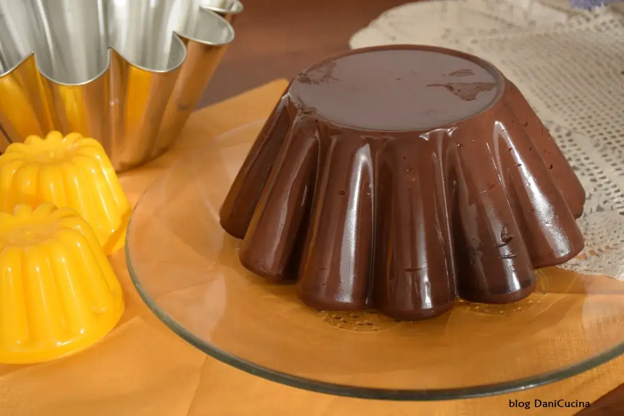 Un lucido budino al cioccolato, appena sformato, riposa su un piatto di vetro. Accanto, due piccoli budini gialli e uno stampo metallico.