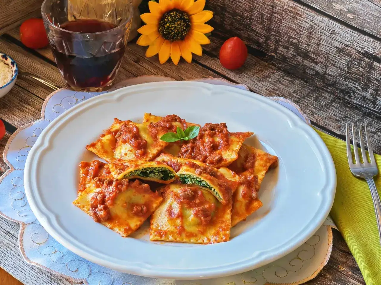 Piatto di ravioli ricotta e spinaci conditi con sugo rosso e rag&ugrave;, guarniti con basilico. Accanto, un bicchiere di vino rosso e un girasole.