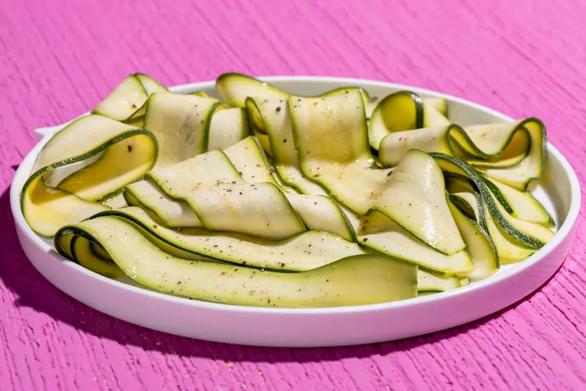 Fettine sottili di zucchine marinate crude, condite con pepe nero, disposte a spirale in un piatto bianco su un tavolo rosa acceso.