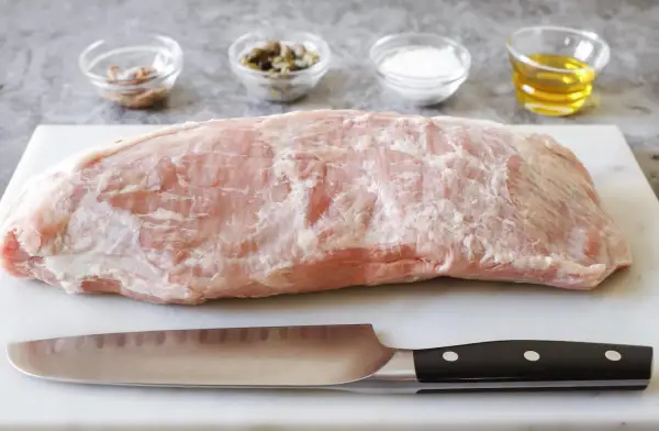 Un pezzo di girello di vitello crudo, pronto per essere cucinato, con coltello e ciotoline di condimenti sullo sfondo.