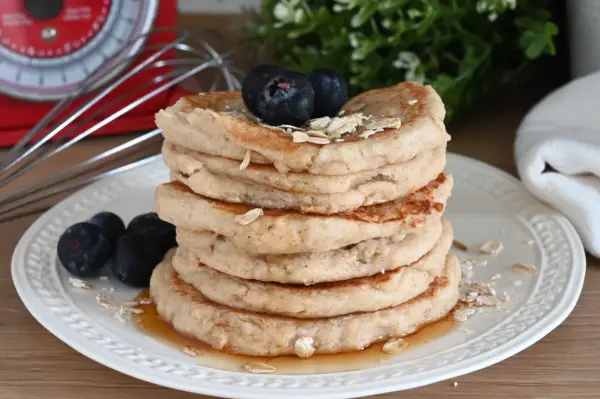 Pila di pancake con farina di avena, guarniti con mirtilli e fiocchi d'avena, su un piatto bianco con sciroppo d'acero.