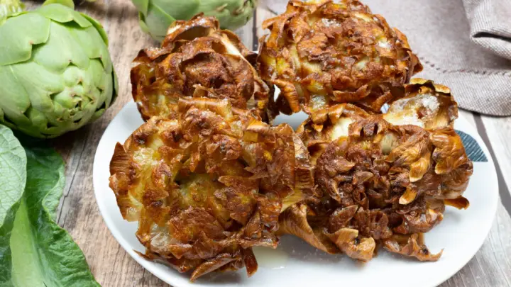 Carciofi alla giudia dorati e croccanti su un piatto bianco, con carciofi freschi sullo sfondo.