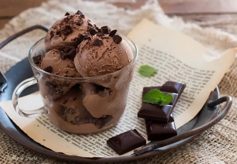 Coppa di gelato al cioccolato con scaglie di cioccolato fondente e foglioline di menta, su un vassoio arrugginito con un libro aperto.