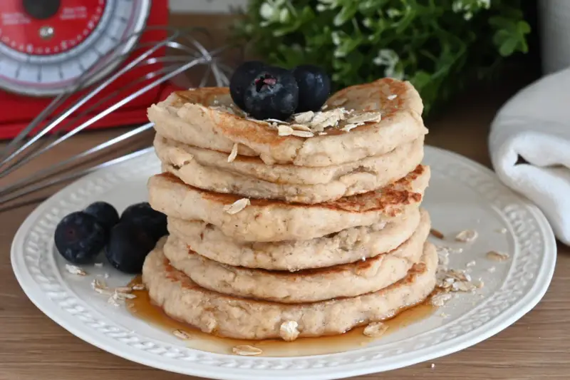 Pila di pancake con farina di avena, guarniti con mirtilli e fiocchi d'avena, su un piatto bianco con sciroppo d'acero.