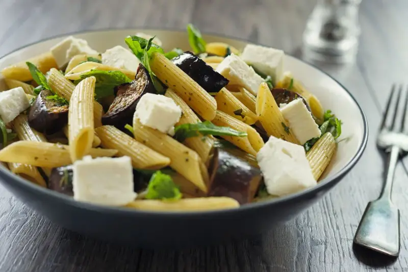 Piatto di pasta con melanzane in bianco, cubetti di formaggio fresco e foglie verdi, servito in una ciotola scura.